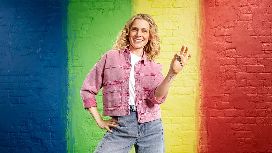 Lachende vrouw in een roze spijkerjasje staat voor een bakstenen muur met verticale kleurvlakken (blauw, groen, geel, rood) en houdt een sleutelbos omhoog.