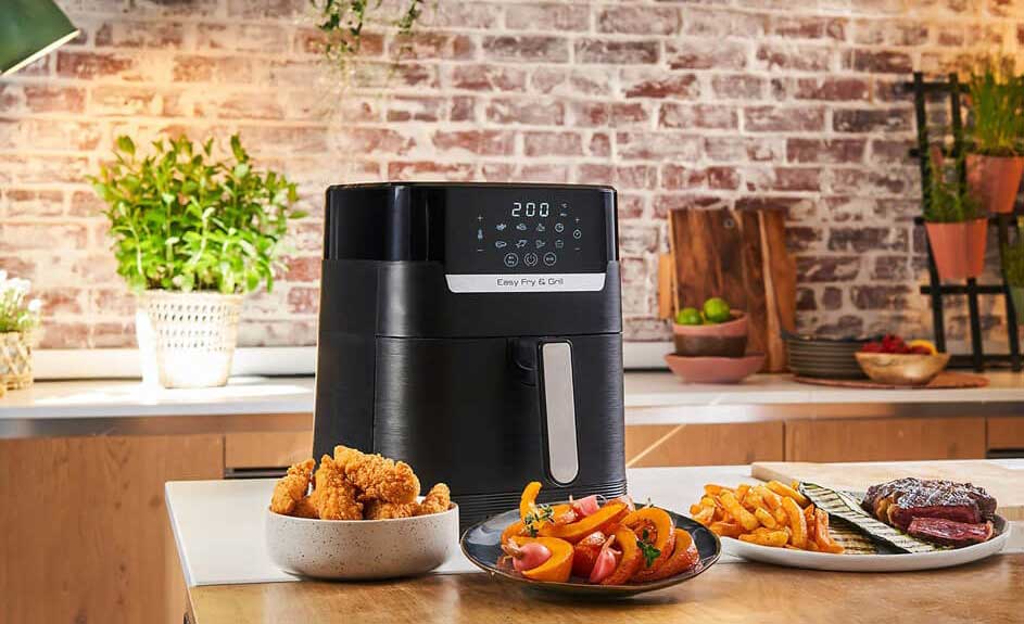Een zwarte airfryer staat op een aanrecht met bereide gerechten, tegen een achtergrond van bakstenen en planten.