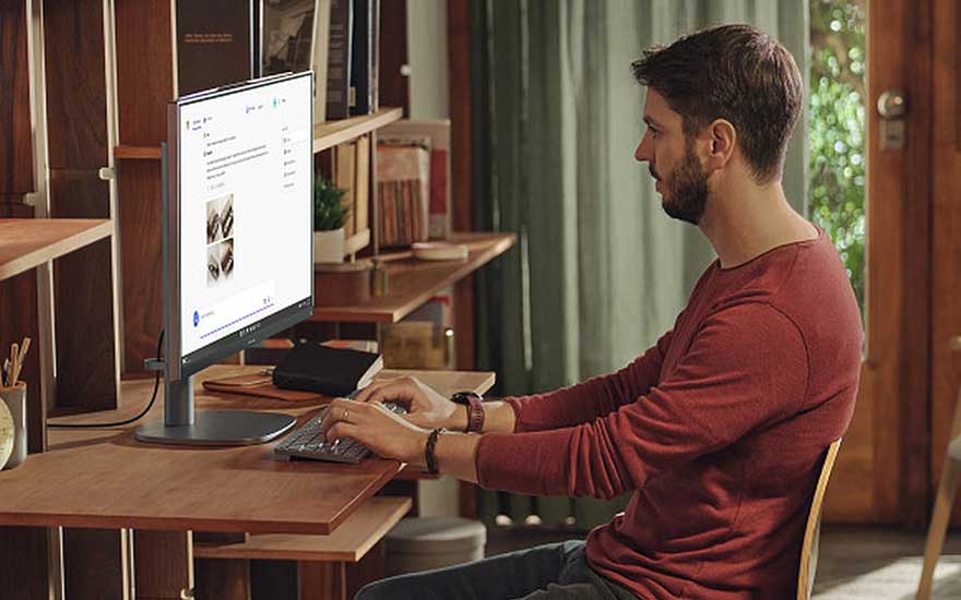 Een man typt aan zijn bureau in een warme thuiswerkomgeving, met een monitor, toetsenbord, muis en houten boekenplanken.