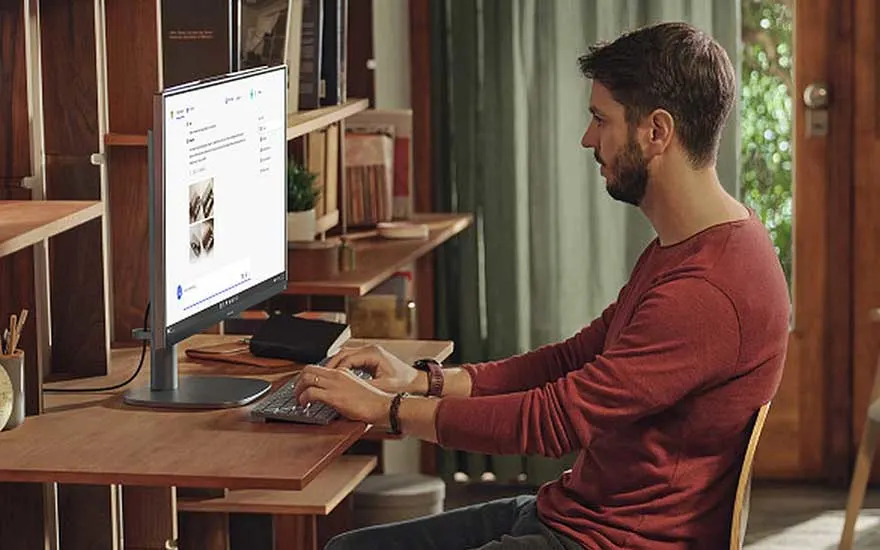 Un homme tape à son bureau dans un espace de télétravail chaleureux, avec un écran, un clavier, une souris et des étagères.