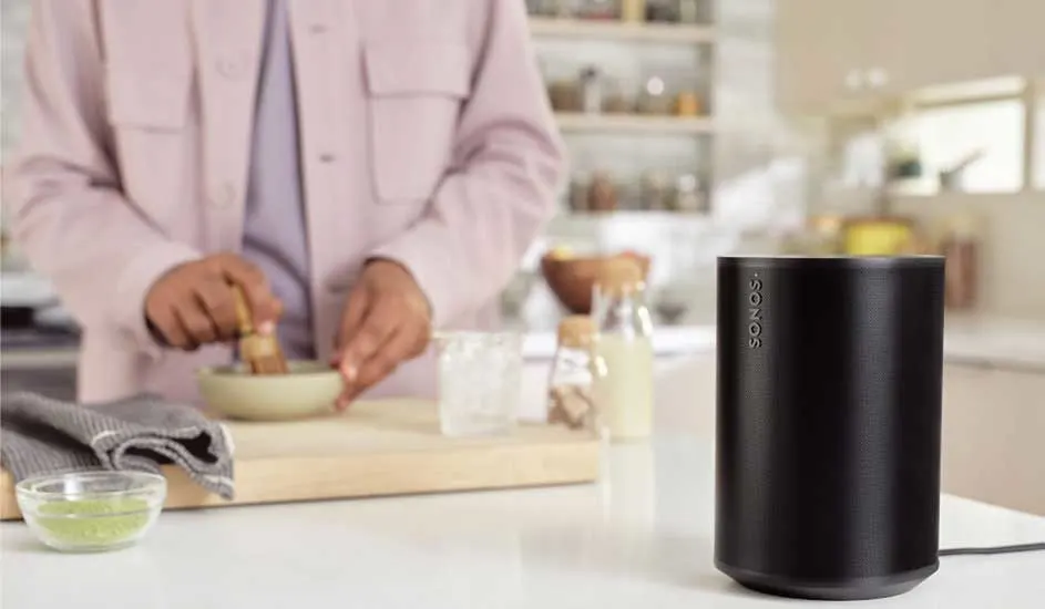 Een zwarte Sonos speaker staat op een witte tafel. Op de achtergrond wordt een matcha drankje bereid in een kom.