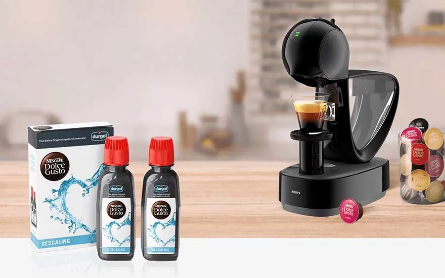 A black Dolce Gusto coffee machine pours espresso, beside descaling bottles, a box and colourful pods on a polished wooden table.
