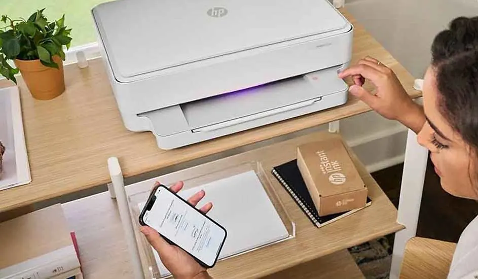 Een witte printer staat op een houten bureau met een potplant en een mobiele telefoon in de buurt.