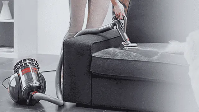 Een grijze stofzuiger met slang staat naast een grijze sofa. De sofa wordt schoongemaakt met een stofzuigermondstuk.