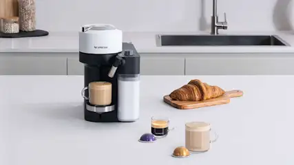 Een zwarte koffiemachine met een witte bovenkant staat op een wit aanrecht met een croissant en koffie.