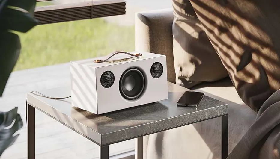 Haut-parleur blanc sur une table grise avec un téléphone portable à côté. En arrière-plan, un canapé et une fenêtre.