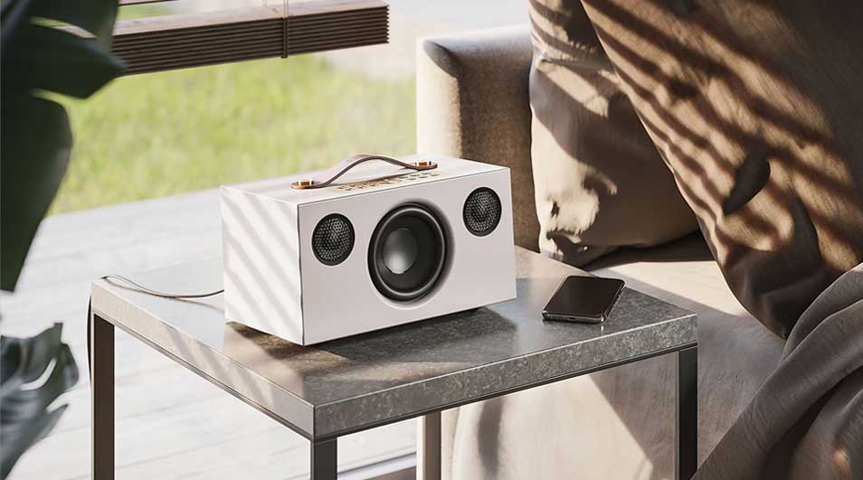 Witte luidspreker met handvat en drie luidsprekers op een grijze tafel. Naast de luidspreker ligt een zwarte smartphone.