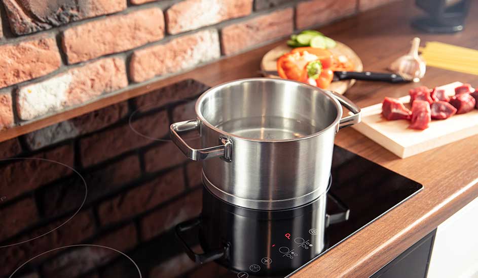 Casserole inox sur plaque de cuisson noire, avec légumes et viande sur plan de travail en bois près d'un mur de briques.