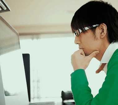 Un écran d'ordinateur blanc est au premier plan, dans un intérieur clair. Une personne est absorbée par l'écran.