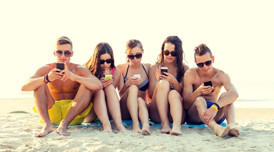 Vijf mensen zitten op het strand en kijken naar hun smartphones. Op de achtergrond is het zandstrand en de zee te zien.