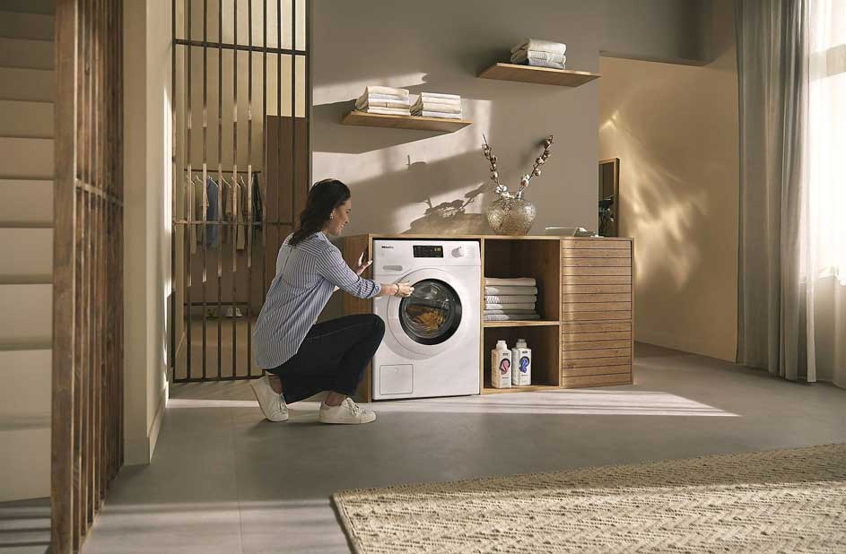 Buanderie : machine à laver blanche à côté d'étagères en bois avec linge et produits. Tapis beige au sol.