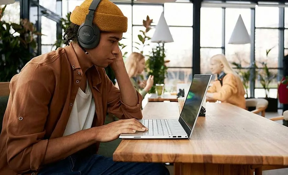 Un homme avec un casque travaille sur un ordinateur portable sur une table en bois. Des plantes et des lumières sont en arrière-plan.