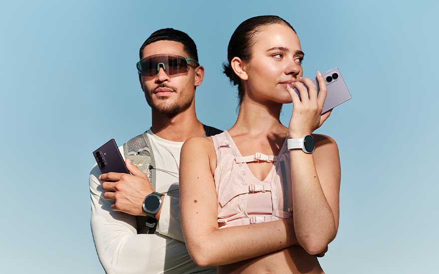 Un couple sportif pose dehors avec des téléphones Samsung et des montres connectées rondes, sous un ciel bleu clair.