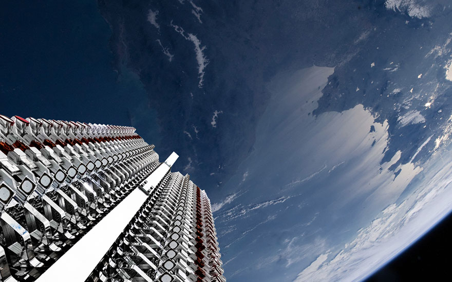Vue plongeante d'une fusée au-dessus de la Terre, avec ciel bleu et nuages. Structures métalliques argentées visibles.