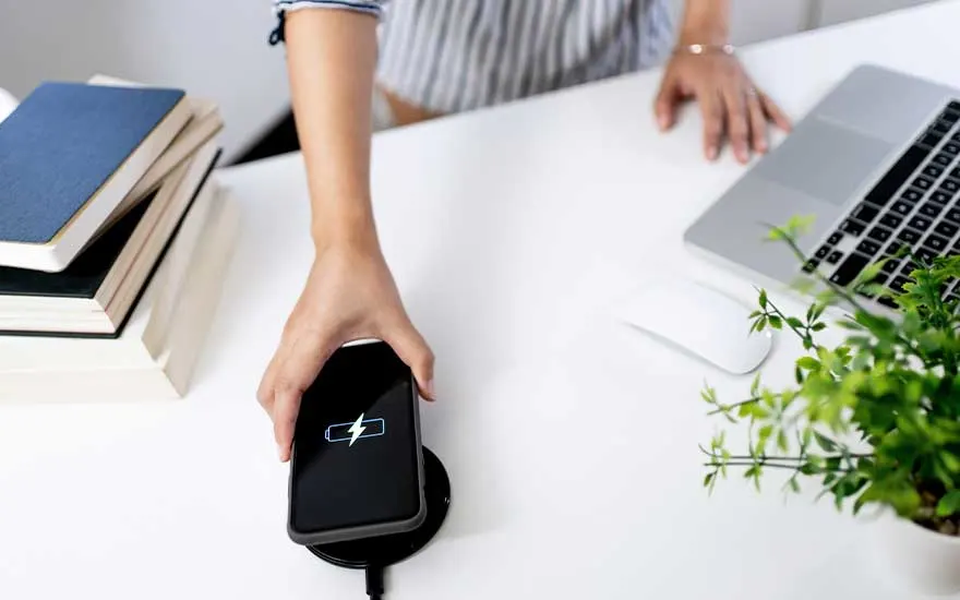 Une main pose un smartphone sur un chargeur sans fil, sur un bureau bien rangé avec ordinateur portable, pile de livres et plante verte.