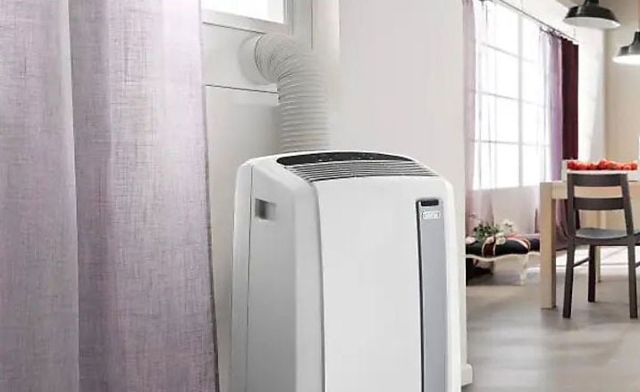 A white portable air conditioner is connected to a window with a hose, near a curtain. A dining table and chairs are in the background.