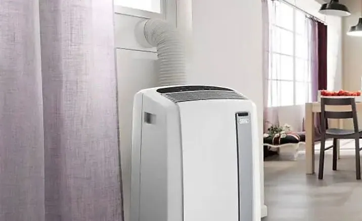 A white portable air conditioner is connected to a window with a hose, near a curtain. A dining table and chairs are in the background.