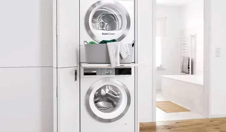 Een Bosch wasdroger staat bovenop een Bosch wasmachine, met een grijze wasmand en handdoek ertussen in een lichte ruimte.