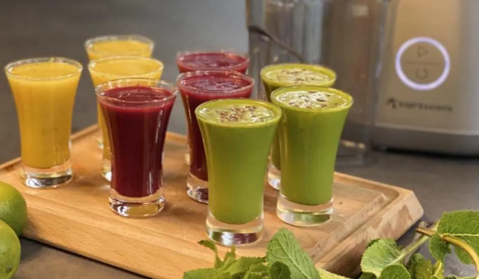 Trois sortes de jus frais dans des petits verres sur un plateau en bois : jaune, rouge et vert, avec une centrifugeuse et des citrons verts derrière.