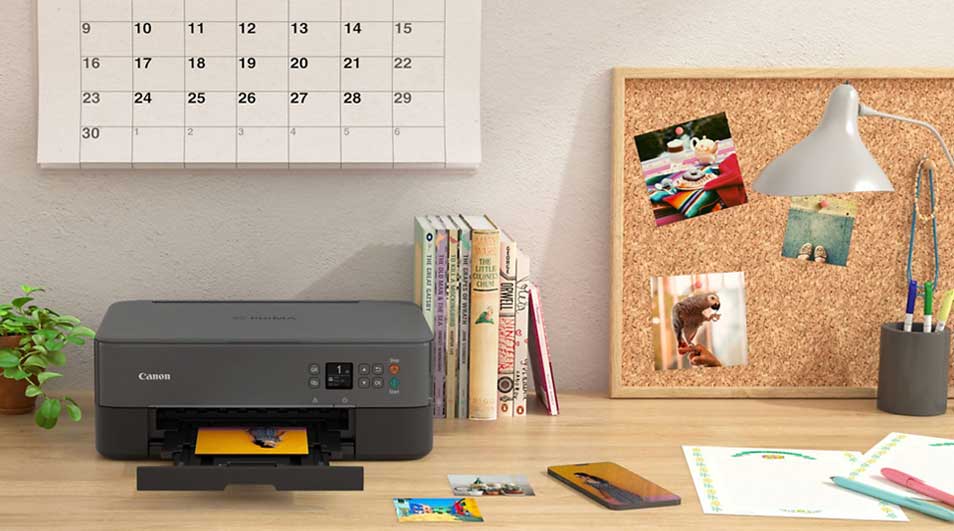 Bureau met printer, kalender, boeken, kurkbord met foto's, lamp, pennenhouder en papier. Kleuren: grijs, bruin, wit, groen.