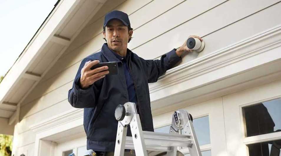 Een witte ladder staat tegen een beige muur, iemand installeert een witte beveiligingscamera en gebruikt een telefoon.