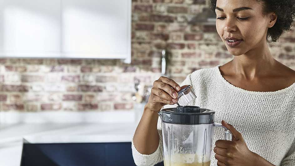 Een blender staat op een aanrecht in een keuken met een bakstenen muur. Iemand giet iets in de blender.