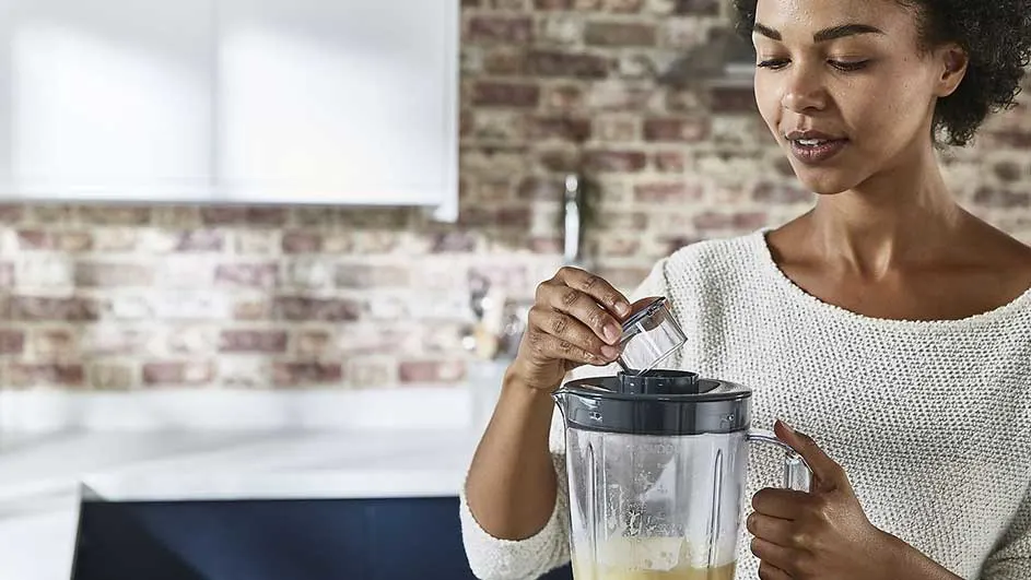 Een blender staat op een aanrecht in een keuken met een bakstenen muur. Iemand giet iets in de blender.