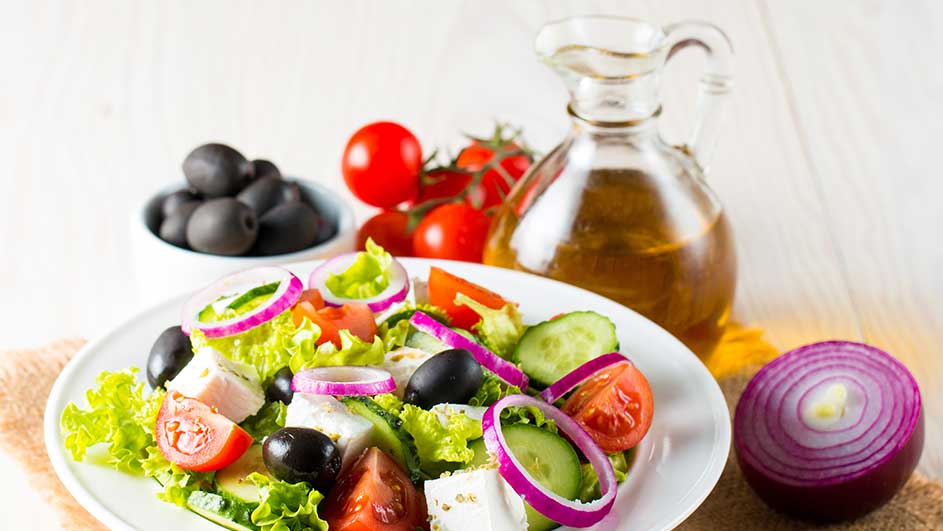 Salade fraîche sur assiette blanche avec tomates, concombres, olives noires, oignons rouges et feta. Huile et olives en arrière-plan.