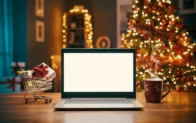 Een laptop met een blanco scherm staat op een houten tafel, met een cadeau in een winkelwagen en een mok ernaast. Kerstboom op achtergrond.