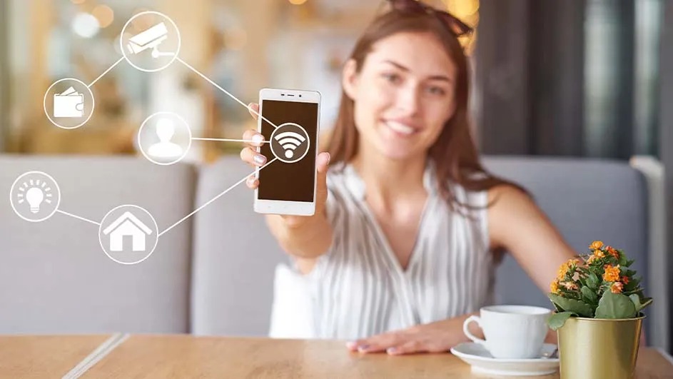 Een smartphone toont een wifi-symbool, verbonden met pictogrammen zoals een huis en een lamp, op een houten tafel met bloemen en een kop.