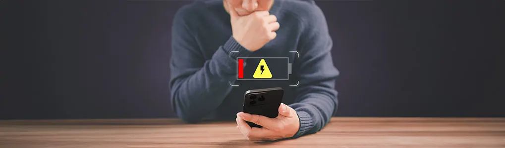 Un smartphone noir affiche une batterie faible avec un éclair jaune sur fond sombre. Table en bois.