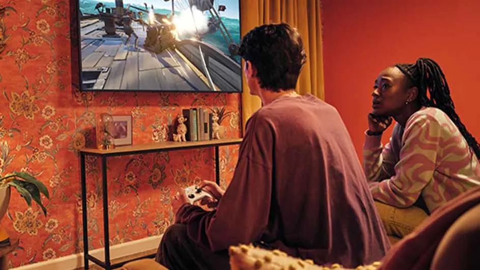 Deux personnes regardent un jeu vidéo sur un écran de télévision dans une pièce aux murs orange à motifs floraux.