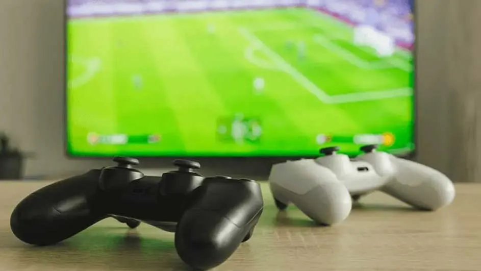 Twee controllers, zwart en wit, liggen op een houten tafel. Een tv-scherm toont een voetbalwedstrijd op de achtergrond.