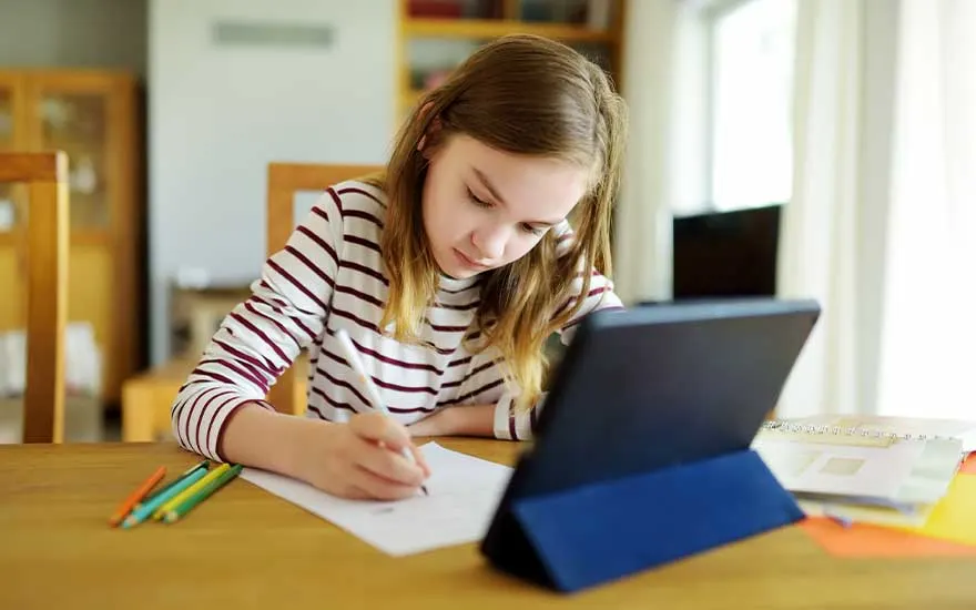 Une tablette noire sur une table en bois, à côté de crayons de couleur et de papiers.