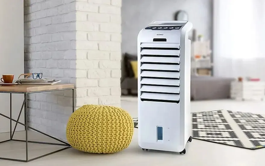 Een witte, verticale ventilator staat naast een gele poef op een vloer met een zwart-wit tapijt.