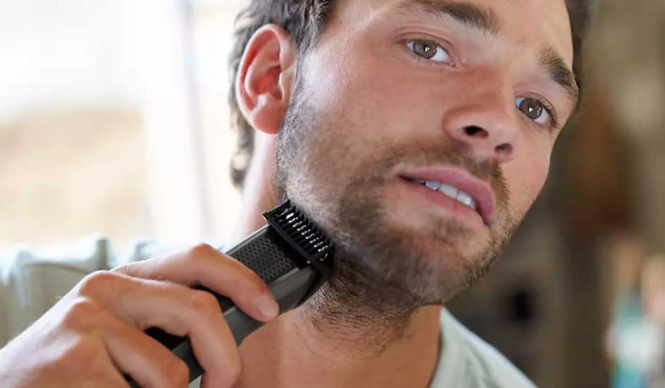 A person is using an electric beard trimmer to trim his beard, carefully shaping the contours of the jawline.