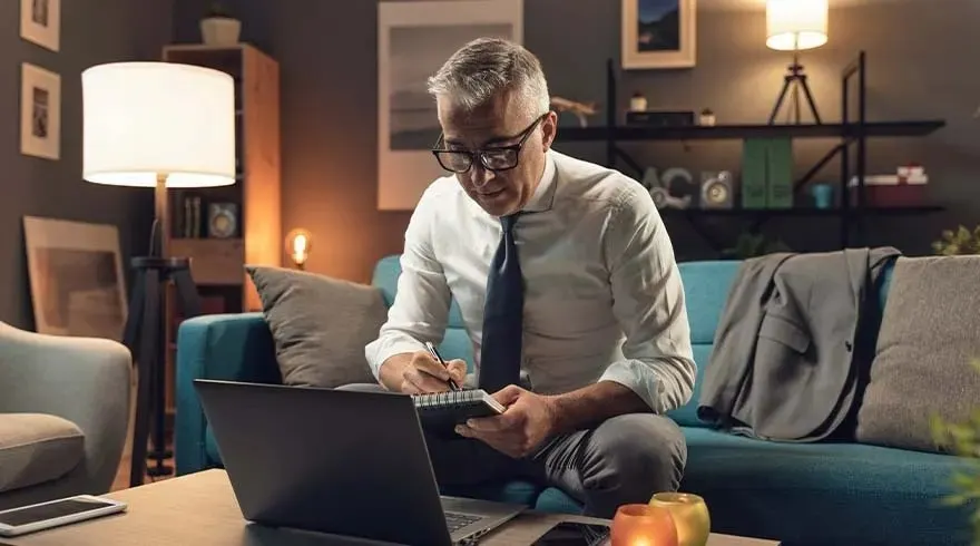 Un homme travaille sur un ordinateur portable et prend des notes sur un canapé bleu dans un salon éclairé.