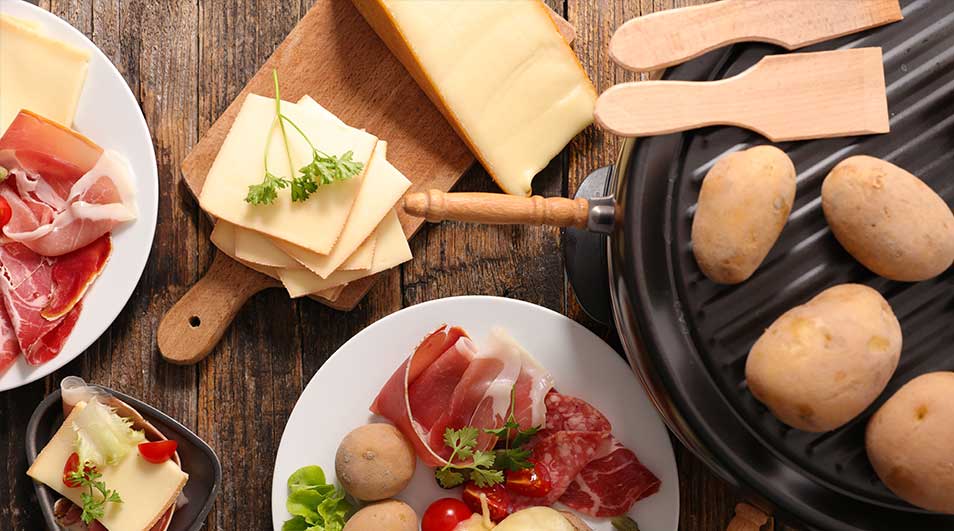 Table en bois avec fromage, charcuterie, pommes de terre et raclette. Assiettes blanches, planches en bois et spatules.