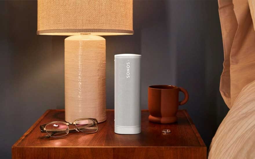 Lampe beige, enceinte Sonos blanche, tasse brune, lunettes et bague sur table de chevet en bois. Fond bleu foncé.