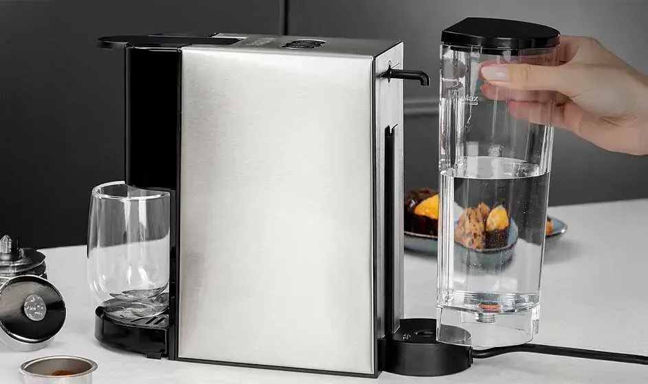 Une machine à café compacte est posée sur un plan de travail, tandis qu’une main remet ou remplit un réservoir d’eau transparent.