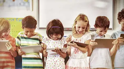 Des enfants alignés utilisent des tablettes dans une salle de classe. Tableau blanc et décorations murales en arrière-plan.