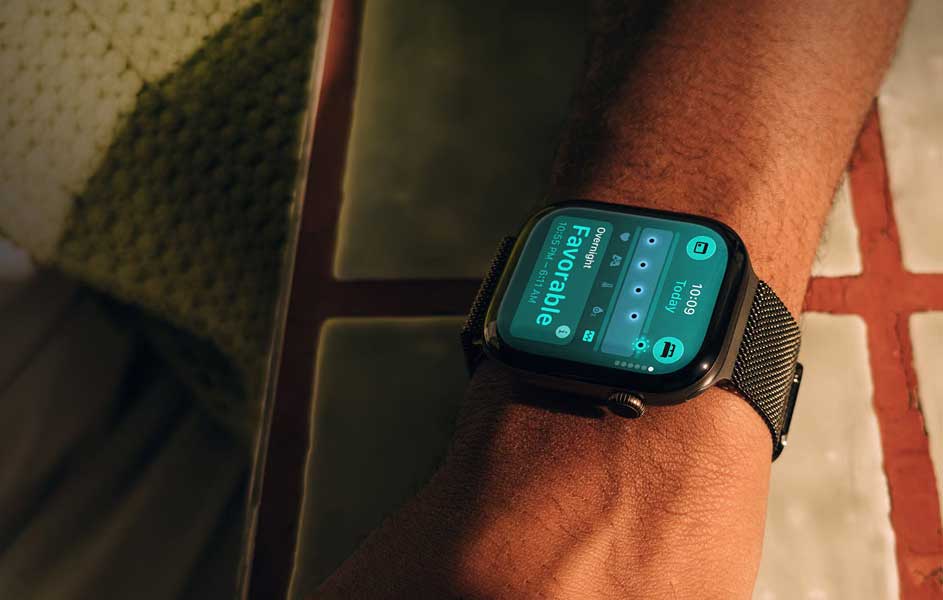 Een persoon draagt een zwarte smartwatch met metalen band. Het scherm toont kleurrijke gezondheidsgegevens, terwijl de arm rust naast een stoel.