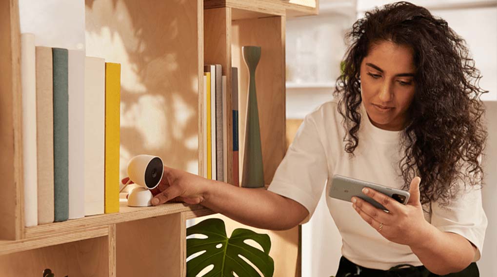 Une femme utilise un smartphone près d'une étagère en bois avec des livres colorés et une petite caméra blanche.