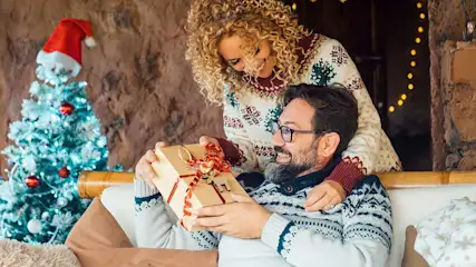 Une femme offre un cadeau joliment emballé à un homme souriant sur le canapé, près d’un sapin de Noël illuminé.