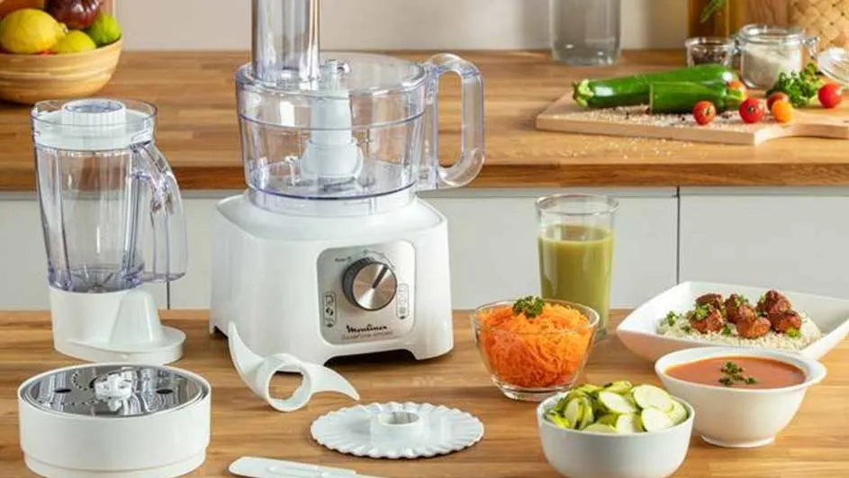 Een witte keukenmachine met transparante opzetstukken staat op een houten aanrecht, omringd door bereide ingrediënten en gerechten.