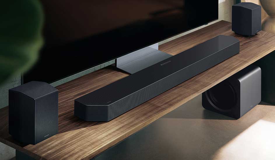 Een Samsung soundbar met twee speakers en een subwoofer staat op een houten tv-meubel onder een grote televisie in een moderne woonkamer.