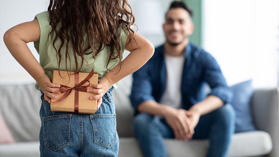 Een meisje houdt een ingepakt cadeau met een bruin lint achter haar rug, met blauwe jeans. Op de achtergrond zit een volwassene op een grijze bank.
