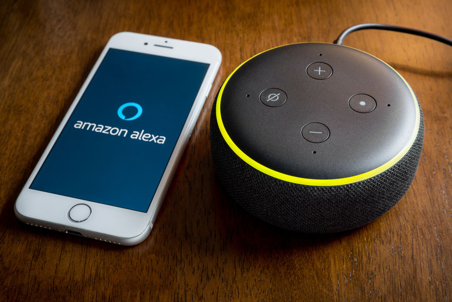 Een witte smartphone met 'amazon alexa' op het scherm, naast een zwarte slimme speaker met gele accenten op een houten tafel.
