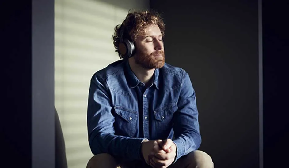 Een persoon met een blauw denim overhemd en een hoofdtelefoon zit met gesloten ogen in een kamer. De achtergrond is donker.