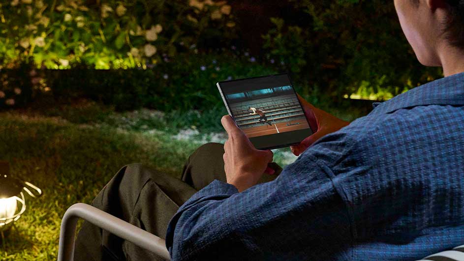 Une personne assise dans un jardin regarde une tablette. L'écran affiche un match de tennis. Des lumières illuminent l'herbe et les plantes.